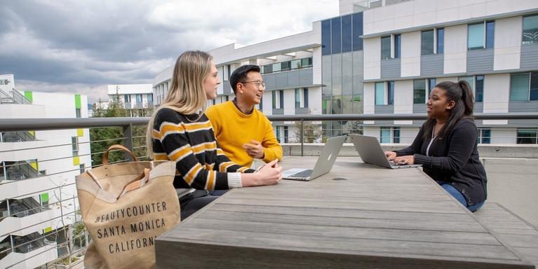 three people talking and working on their laptops while sitting together on a table on a rooftop