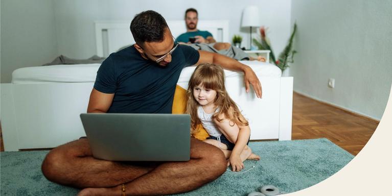 parent sitting cross-legged on a rug in front of a bed with a laptop on their lap and child next to them, with their partner visible in the background