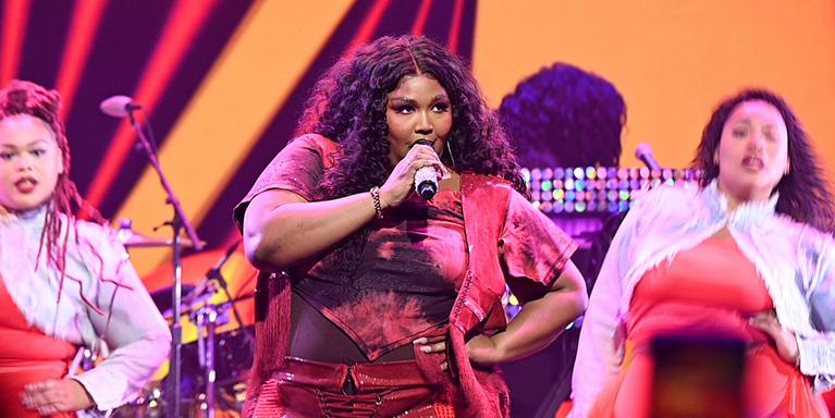 Lizzo performs onstage in front of two dancers and an orange, red, and purple backdrop