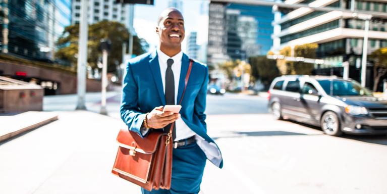 person in blue business suit with brown leather bag walking through city, looking up from phone and smiling