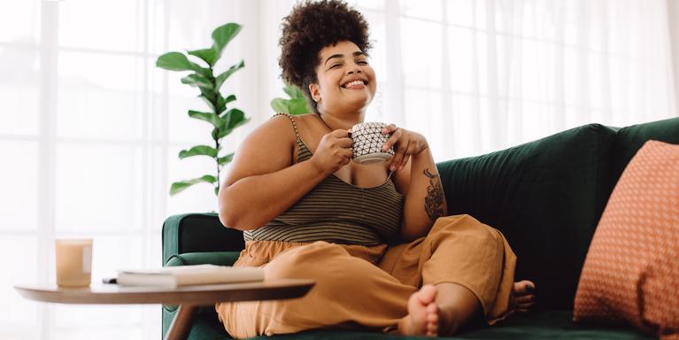 person sitting cross-legged on dark green couch, holding coffee mug and smiling