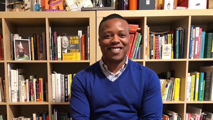 A man in a blue sweater smiling in front of a bookshelf