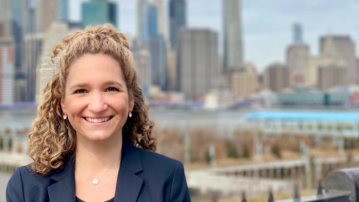 a person with light-colored curly hair, wearing a blazer and smiling in front of a city skyline