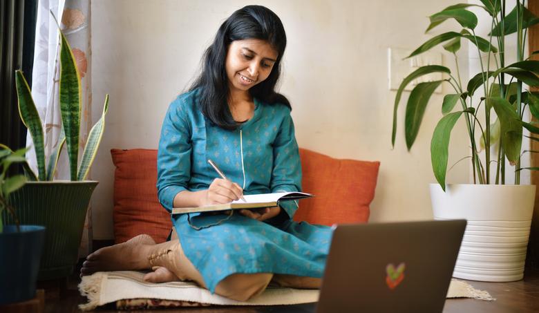 person kneeling on floor in front of orange pillows, writing in a notebook while looking at open laptop