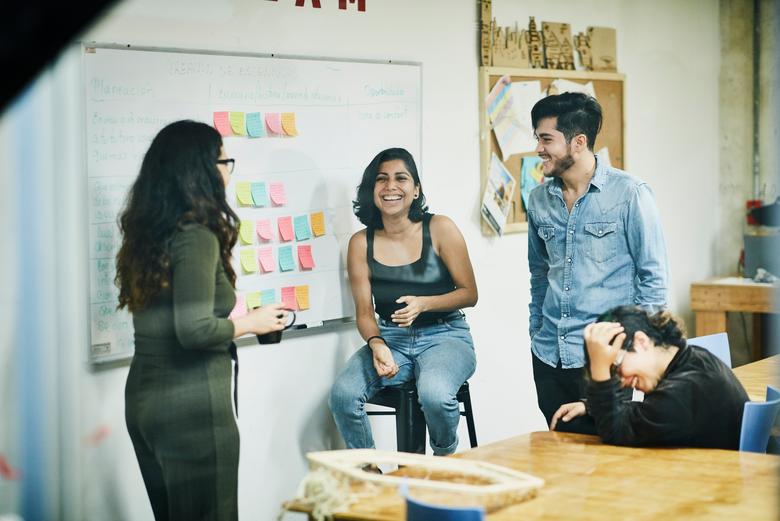 a group of coworkers sitting and standing in front of a white board with colorful sticky notes on it, talking and smiling