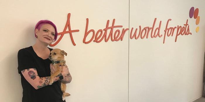 person with short pink hair holding a dog and standing against a wall that says "A better world for pets."