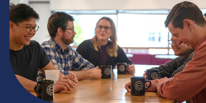 A group of smiling people gathered at a conference table. Each person has a black coffee mug in front of them.