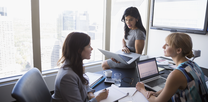 three people sitting at an office table with laptops and notepads