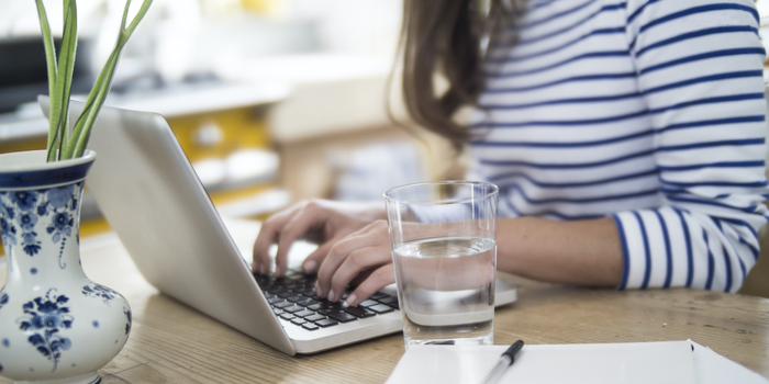 person sitting at a table typing on a laptop with a glass of water and notebook and pen nearby