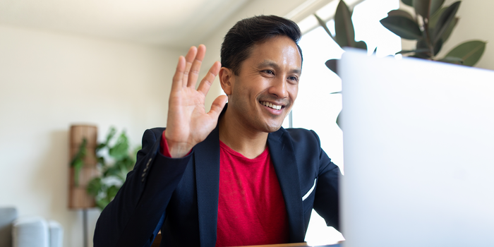 person sitting at home waving hello while on a video call on their laptop