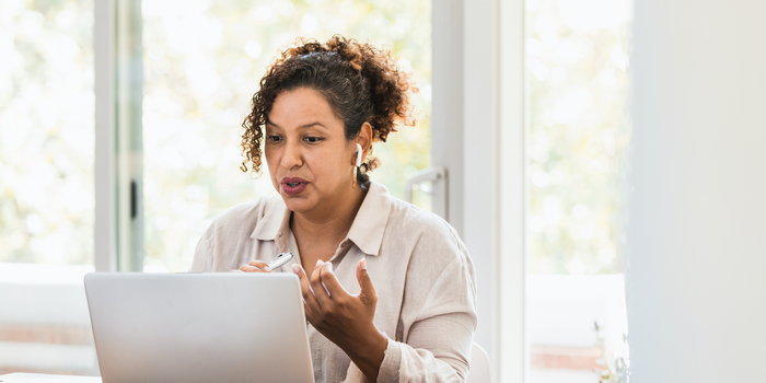 person sitting at home in front of a laptop wearing earbuds and speaking while in a remote meeting