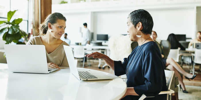 two coworkers sitting at a table with laptops open having a conversation