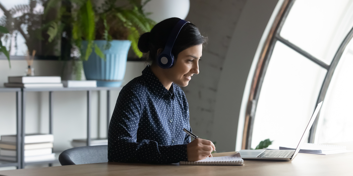 person sitting at a table at home looking at their laptop, wearing headphones, and taking notes in a notebook