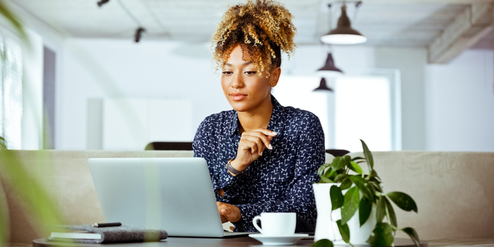 person sitting at a table looking at their laptop with a notebook, coffee cup, and plant nearby