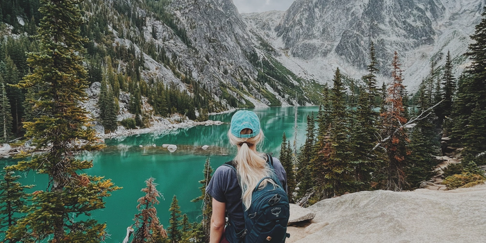author April Zimmerman stands with her back to the camera looking out on a lake between mountains studded with trees, wearing a backpack and her hair in a ponytail under a baseball cap
