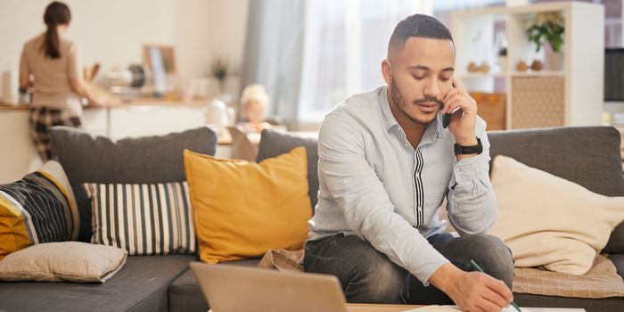 Person sitting on their couch at home with an open laptop in front of them, talking on the phone and taking notes.