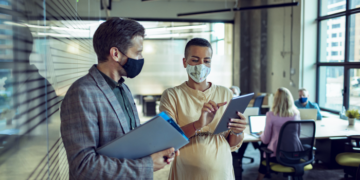 two people wearing masks standing and talking in an office, one holding and pointing to a tablet, the other holding folders