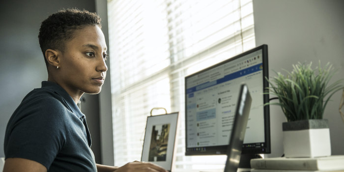 person sitting at their desk at home with a computer monitor and laptop open, looking at the latter