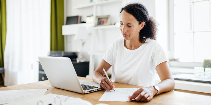 person sitting at a table at home looking at an open laptop and taking notes on a piece of paper