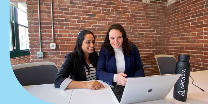 Two people sitting at a conference table and looking at a laptop. They are smiling; a brick wall can be seen in the background.
