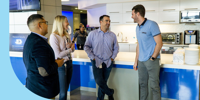 Four people dressed in business casual attire and engaged in a conversation in an office area.