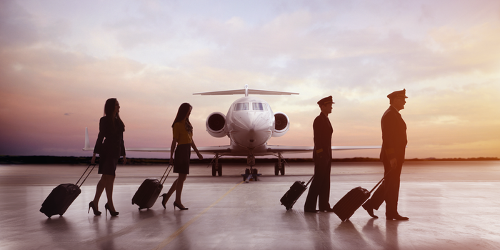 four members of a flight crew wheel their bags across the tarmac in silhouette with the nose of a plane in the background between them