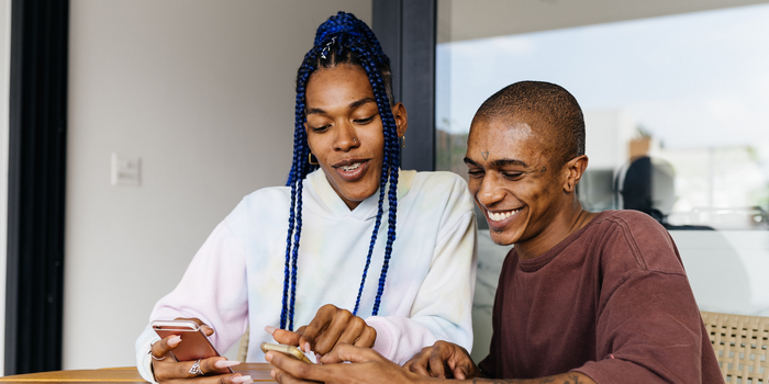 two coworkers sitting at a conference table holding cell phones pointing to one of their screens and smiling