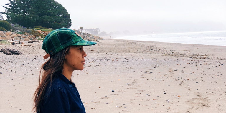 photo of the author, Chante Owens, wearing a cap, standing on the sand at the beach, looking out at the water, with a few trees visible on the shoreline behind her