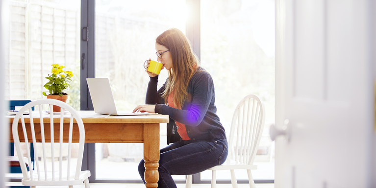 person sitting at their kitchen table, drinking from a mug, and working on a laptop