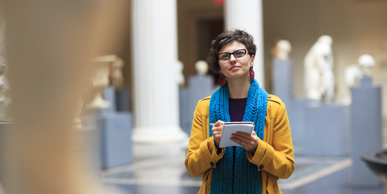 young adult standing in a museum looking at sculptures (others are visible in the background) and taking notes in a small notebook