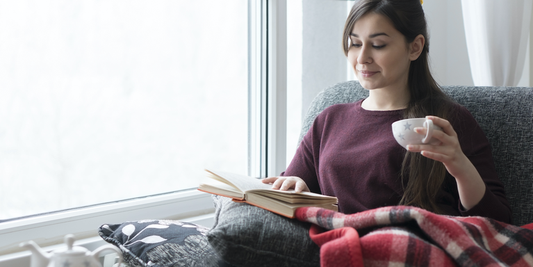 person sitting on a couch reading a book and drinking tea with a blanket on their lap