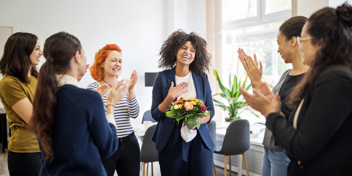 smiling person standing in an office holding flowers in one hand and touching the other to their chest in gratitude as their colleagues stand around them and clap